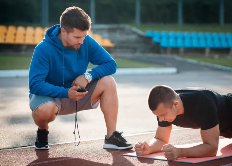 Un uomo con una felpa blu con cappuccio si accovaccia con un cronometro, cronometrando un altro uomo con una camicia nera che sta tenendo una posizione di plank su un tappetino per esercizi in una pista all'aperto.