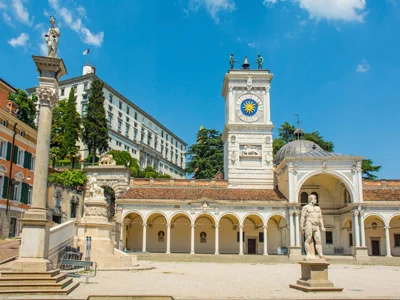 Una piazza soleggiata con una torre dell'orologio bianca, un colonnato ad archi, statue di marmo ed edifici storici circondati dal verde in un cielo blu brillante.